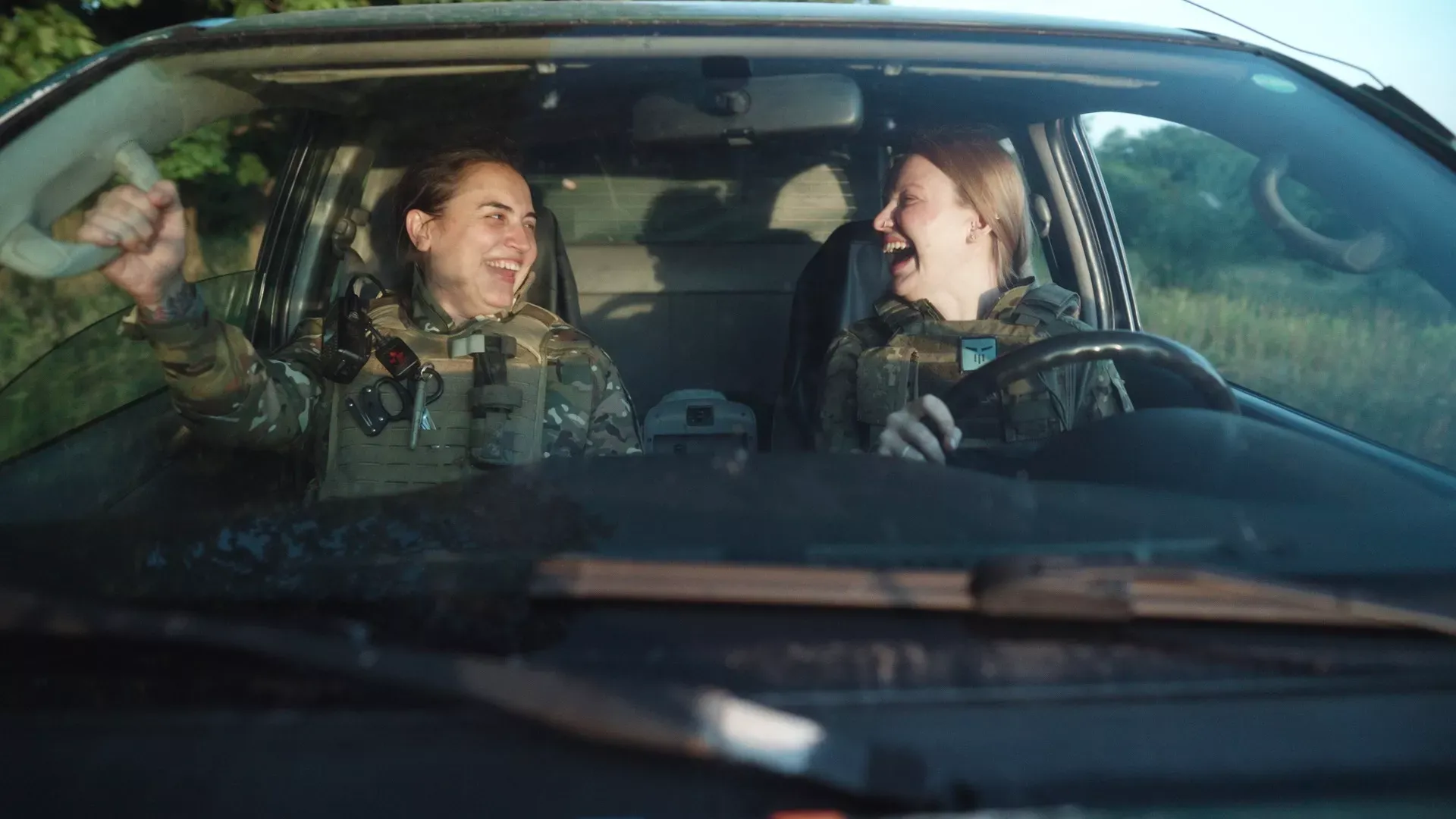 Deux femmes en tenue militaire sont dans une voiture. Elles se regardent et rient.